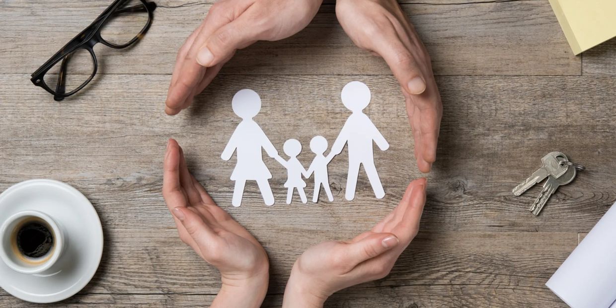 Hands protect paper cutout family figures on a wooden table.