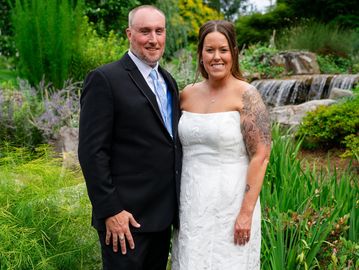 A couple in wedding attire posing outdoors near a waterfall.