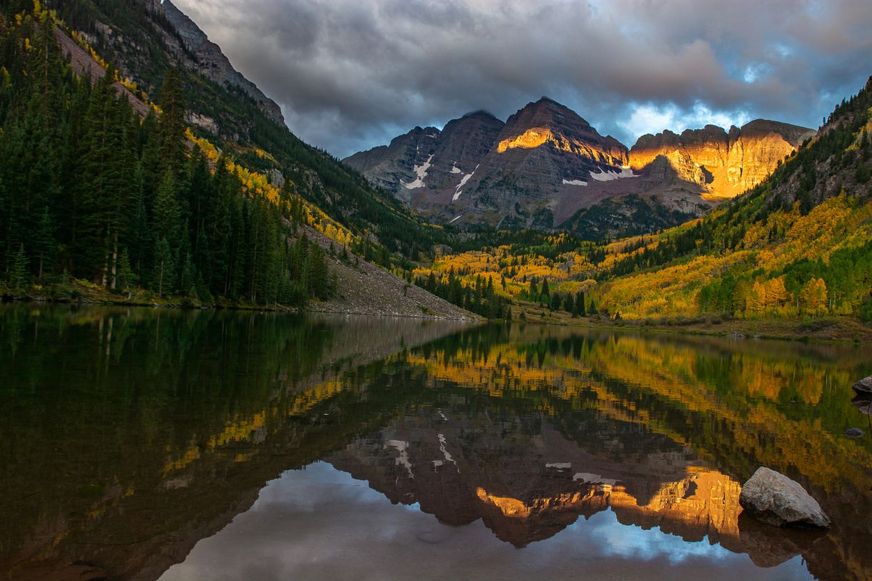 Sunlit mountain peaks reflecting on a calm lake surrounded by autumn trees.
