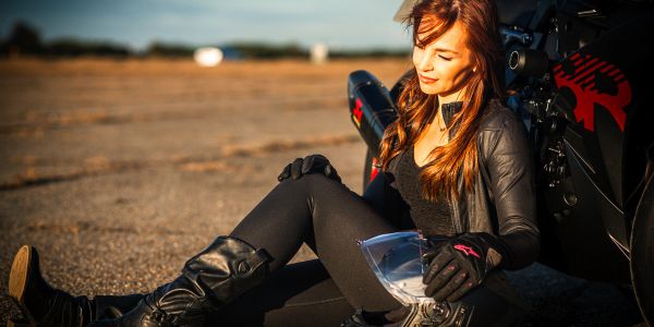 Woman in motorcycle gear sitting by her bike at sunset.