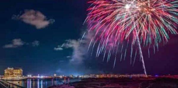Colorful fireworks explode over a city waterfront at night.