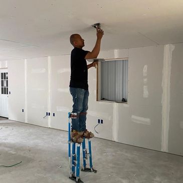 Worker sanding ceiling drywall while standing on stilts in unfinished room.