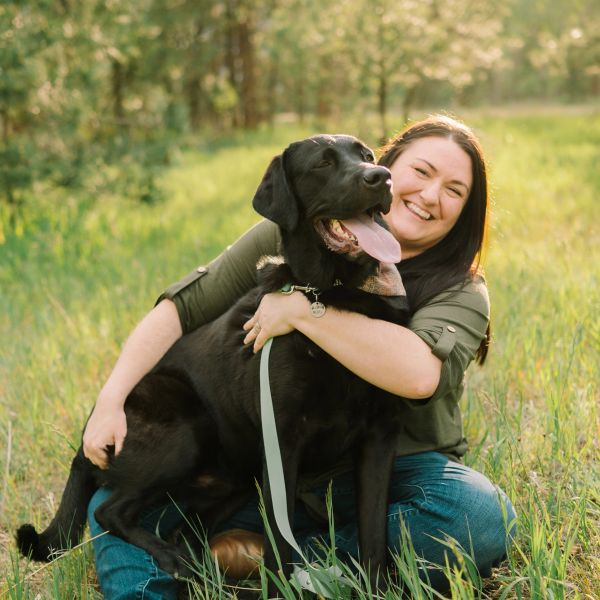 Woman happily embraces a large black dog in a sunny meadow.