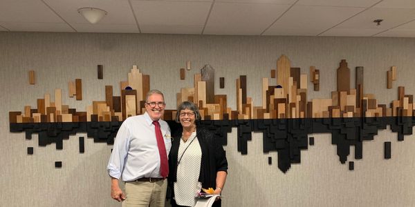 Two people posing in front of a wooden city skyline wall art.