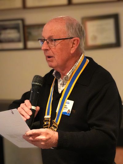 Elderly man speaking into a microphone, holding papers, wearing a medal and name tag.