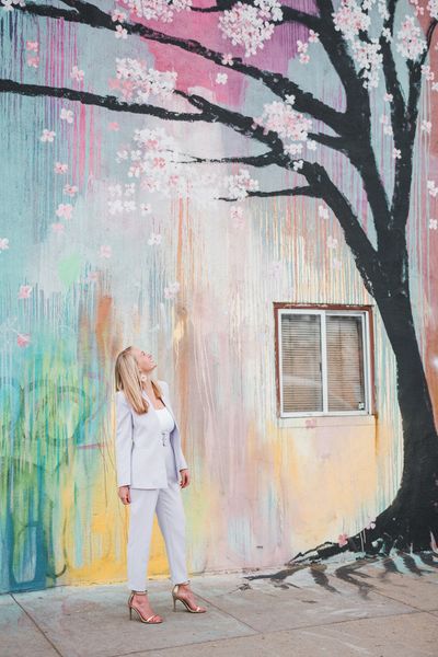 a blond woman in a business suit stands in front of a mural looking up at a tree painted on building