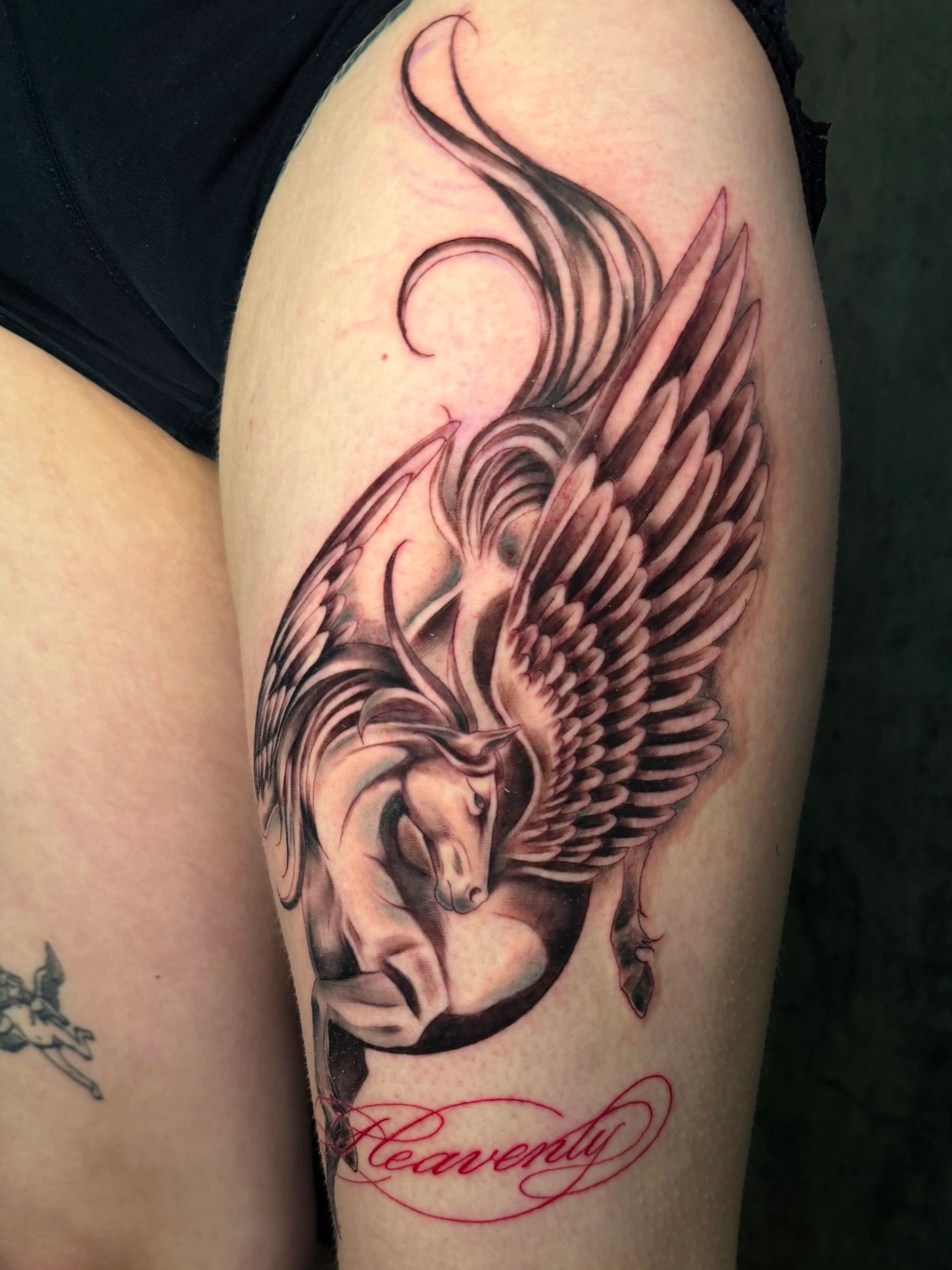 Black and grey tattoo of a winged horse with red script.
