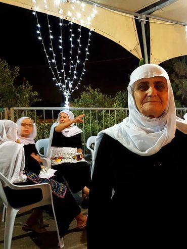 Elderly woman in white headscarf among seated women with festive lights.