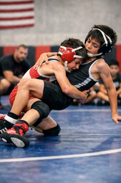 Two youth male wrestlers compete on a gym mat, with one in a red singlet executing a takedown