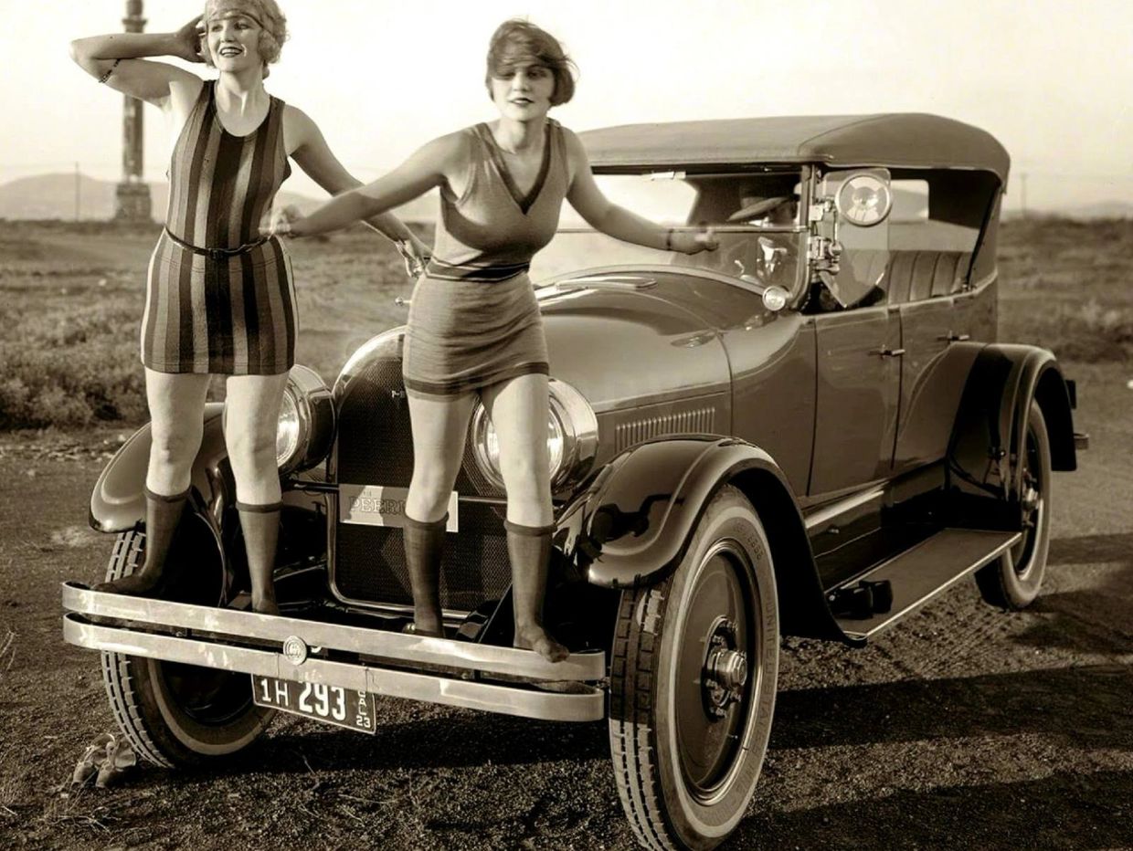 Two women in vintage swimwear balancing on an old car's bumper.