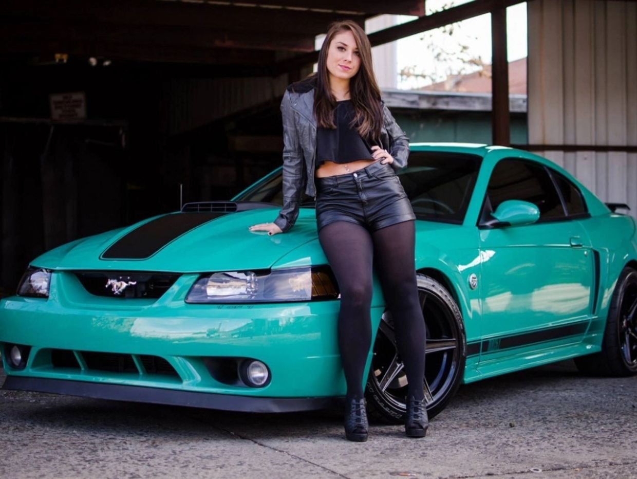 Woman leaning on a teal sports car under a covered area.