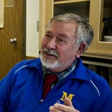 Older man in a blue jacket speaking indoors.