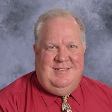 Smiling man in a red shirt and patterned tie against a gray background.