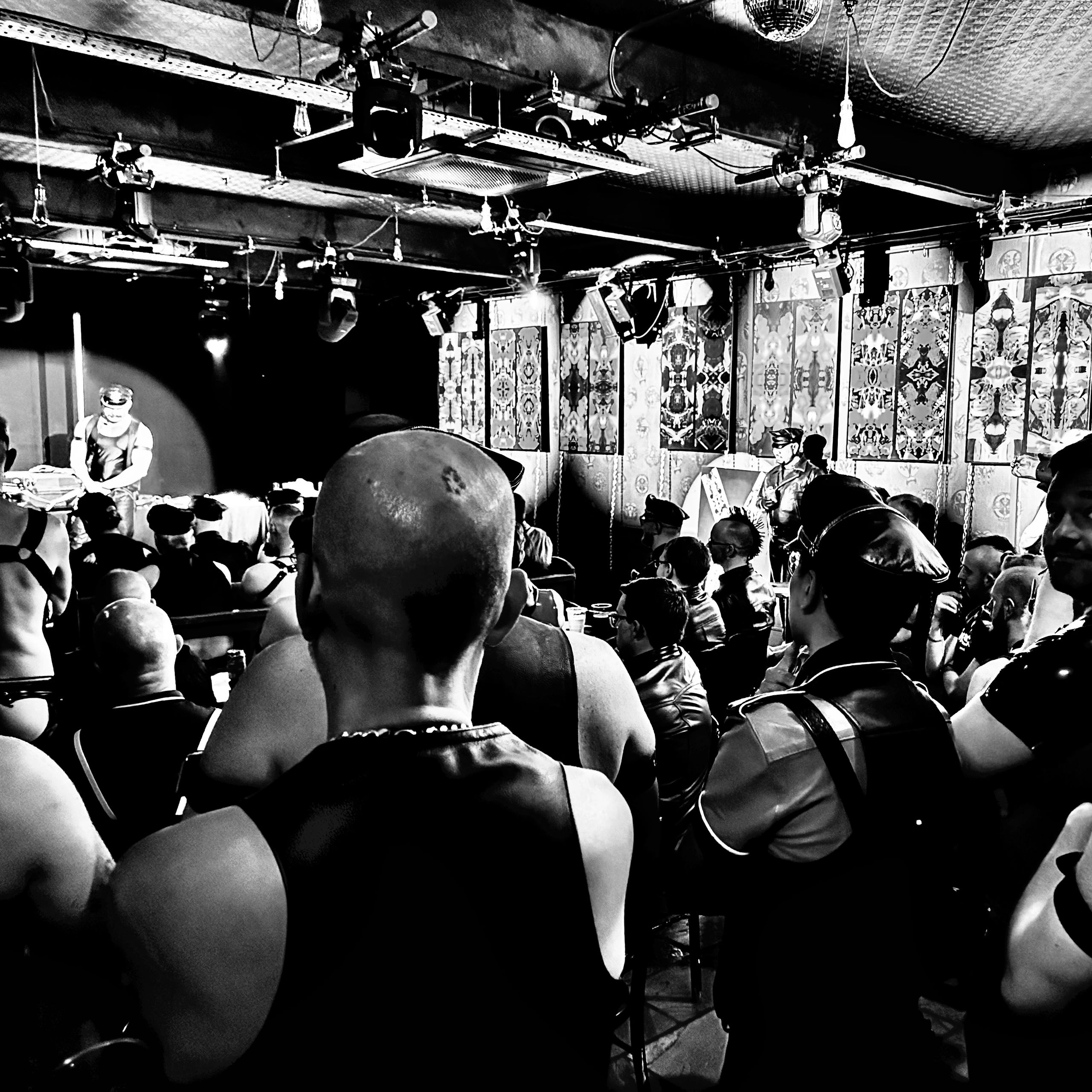 Black and white scene of a crowded club with many people in leather outfits and a DJ.