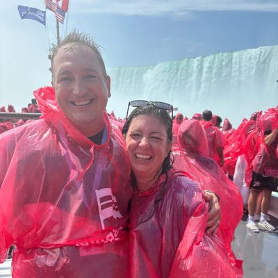 Two people in red ponchos smiling near a waterfall with a crowd behind them.