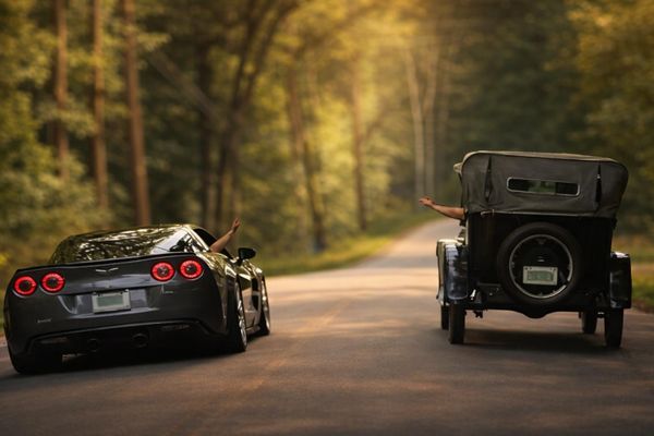 A modern sports car and a vintage car driving side by side on a forest road.