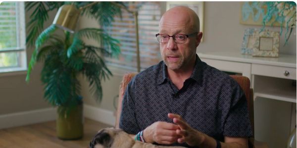 Bald man wearing glasses sits indoors with a pug on his lap.