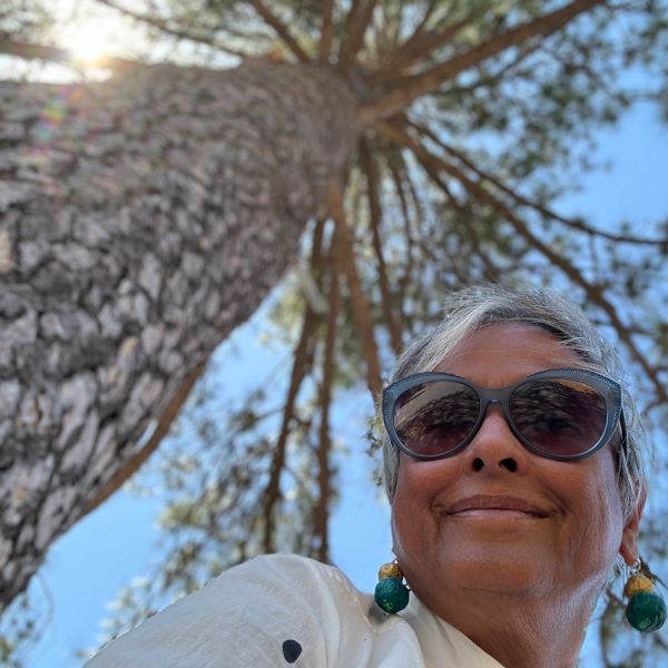A woman wearing sunglasses smiles under a tall tree on a sunny day.