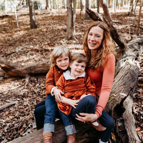A smiling woman and two children sitting on a fallen tree in a forest.