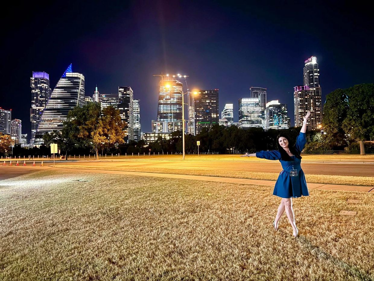 Woman in blue dress posing joyfully in front of a lit city skyline at night.