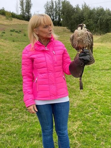 A woman in a pink jacket holding a falcon on her gloved hand outdoors.