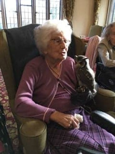 Elderly woman sitting with a small owl on her gloved hand indoors.