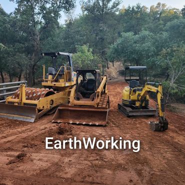 Three yellow earthworking machines on a dirt path in a forested area.