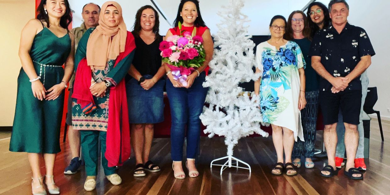 A diverse group of nine people standing indoors around a white Christmas tree, smiling.