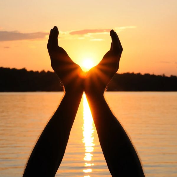 Hands framing the glowing sun at sunset over a calm lake.