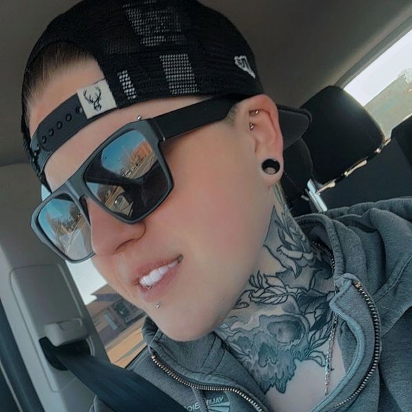 Person with sunglasses, tattoos, and piercings sitting in a car.