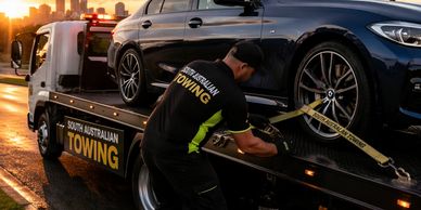 A worker secures a black car on a South Australian towing truck at sunset.
