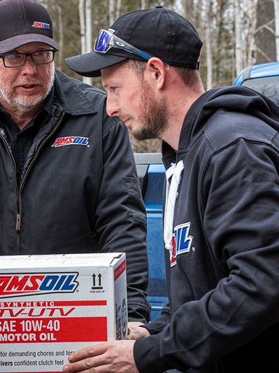 Two men in black jackets with Amsoil logo load AMSOIL  products into the back of a truck.