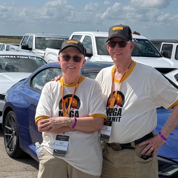 Two smiling people wearing matching shirts and hats at a car event.