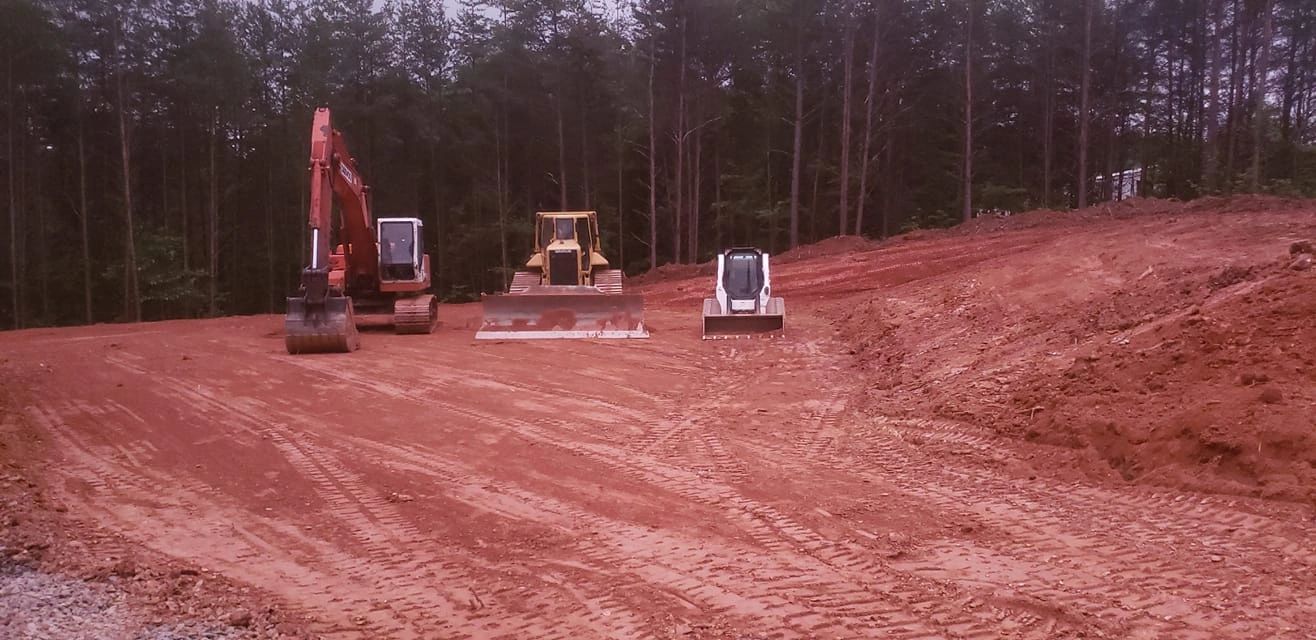 Three construction machines parked on a cleared dirt area near a forest.