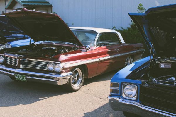 Two classic cars with open hoods at a car show on a sunny day.