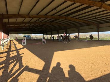 Covered equestrian arena with riders on horses practicing jumps.