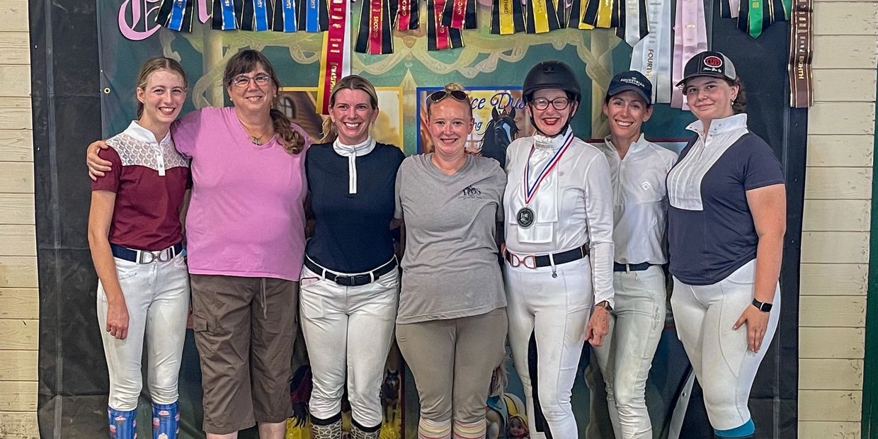 Group of seven women standing in front of a wall decorated with numerous colorful award ribbons.