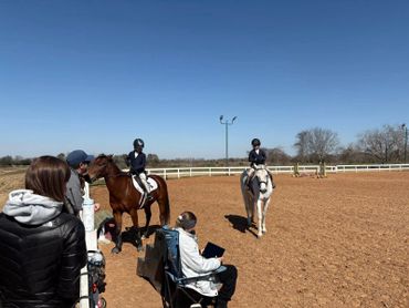 Children dressed for horseback riding in an outdoor arena with spectators.