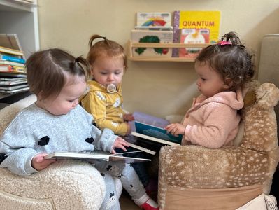 Three toddlers sitting on plush chairs, reading books together in a cozy room.