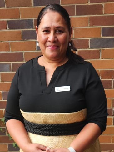 Woman in traditional attire standing against a brick wall, smiling warmly.