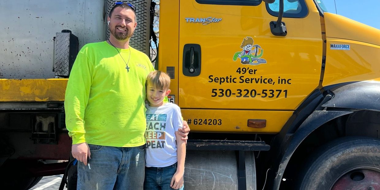 Man and boy standing by a yellow septic service truck on a sunny day.