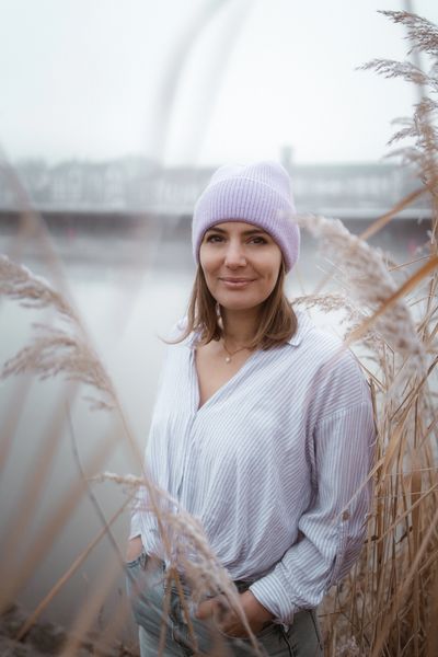 Young woman in a lilac beanie smiling by the water with tall grasses around her.
