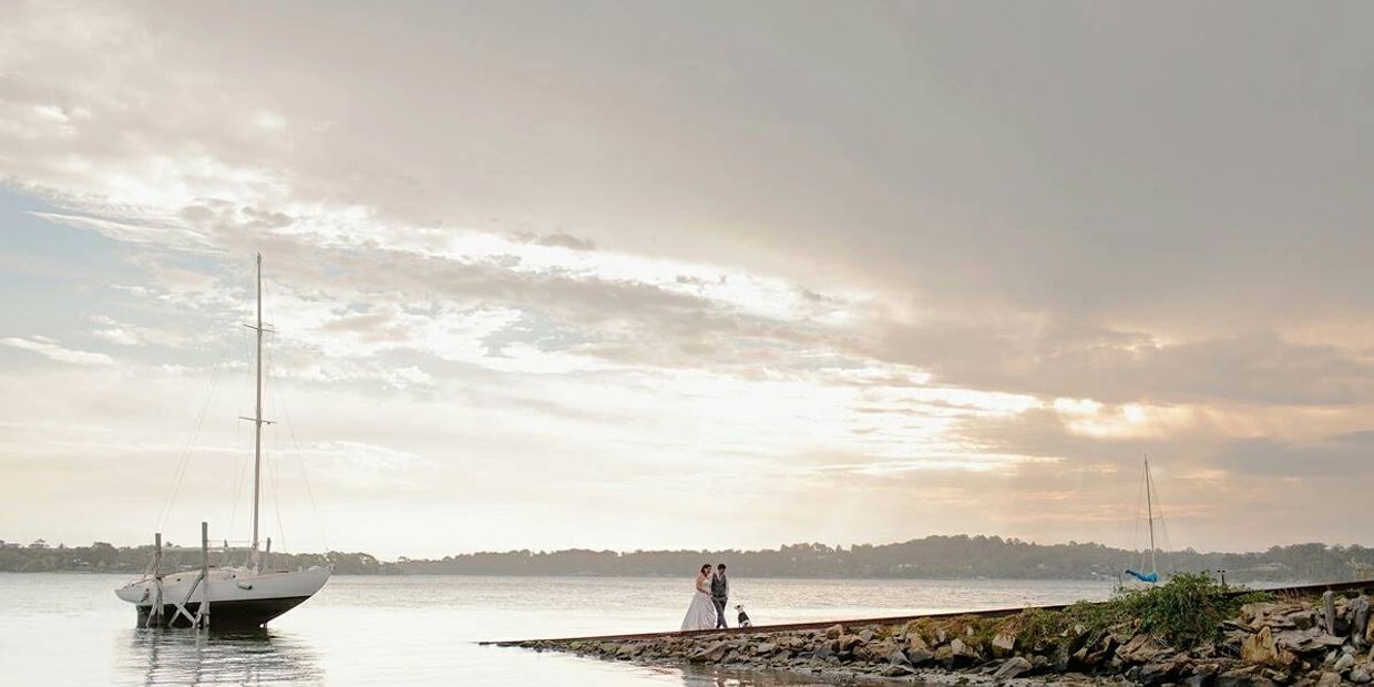 A bride, groom and dog stand on a slipway at sunset, a yacht is on a cradle in the water behind