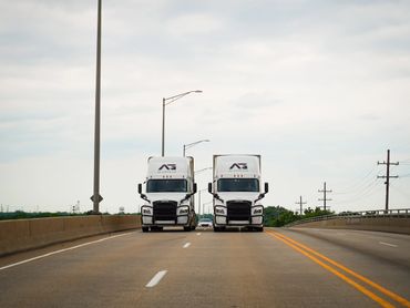 Two white semi-trucks driving side by side on a highway under a cloudy sky.