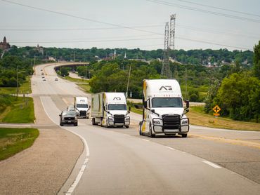 Three white semi trucks and a car driving on a highway surrounded by greenery.