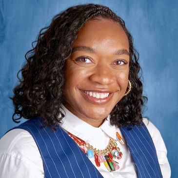 Smiling woman with curly hair wearing a pinstripe vest and colorful necklace.