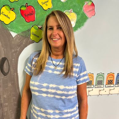 A smiling woman stands in front of a colorful classroom wall with apple-shaped name tags.