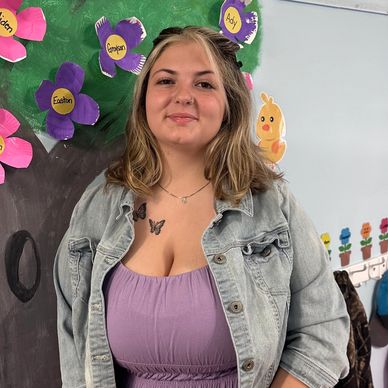Young woman with butterfly tattoo wearing lavender dress and denim jacket in a colorful classroom.
