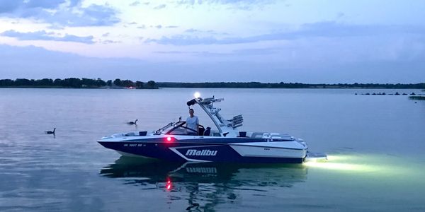 A Malibu boat floats on calm water at dusk with lights on.