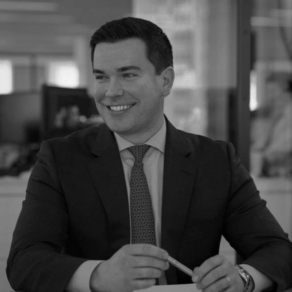 Smiling man in a suit holding a pen, seated indoors.